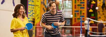 Two adults hold blue table tennis paddles, rallying and smiling, in an indoor recreation center; a white ball and colorful climbing walls and equipment are visible in the background.