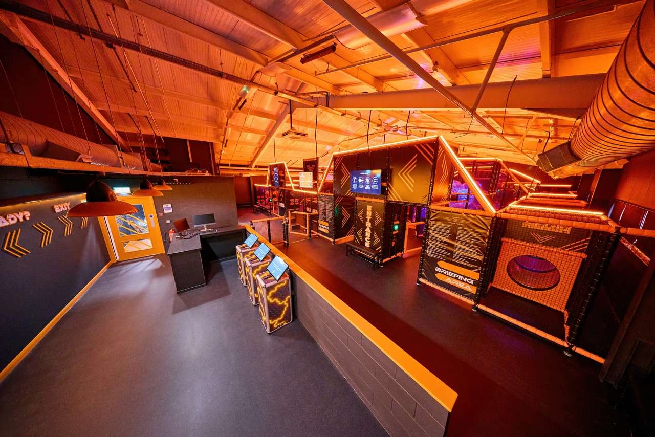 Neon-framed indoor obstacle course glows, guiding entrants toward gated sections; touch-screen kiosks await check-in under an industrial ceiling. Visible text: READY?, EXIT, ENTRANCE, BRIEFING AREA.