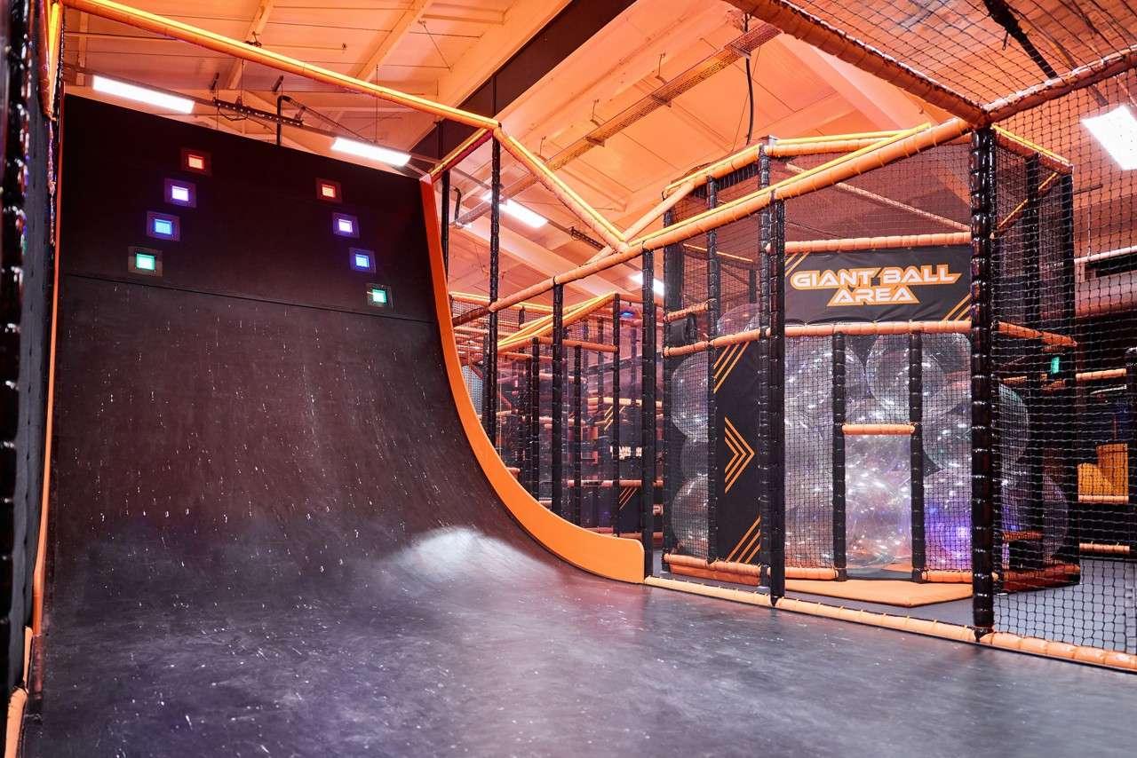 Curved ramp rises steeply with illuminated square targets, beside a netted indoor play structure labeled GIANT BALL AREA containing large transparent balls under orange-framed beams.