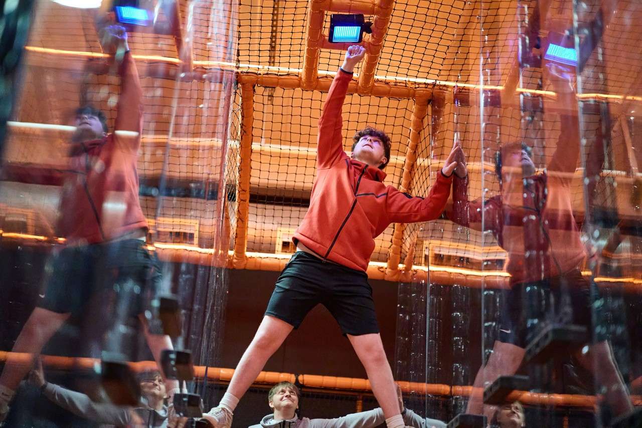 Boy in red jacket presses a blue button while climbing between transparent walls; safety netting and orange tubing enclose an indoor obstacle course, as a child watches and supports below.