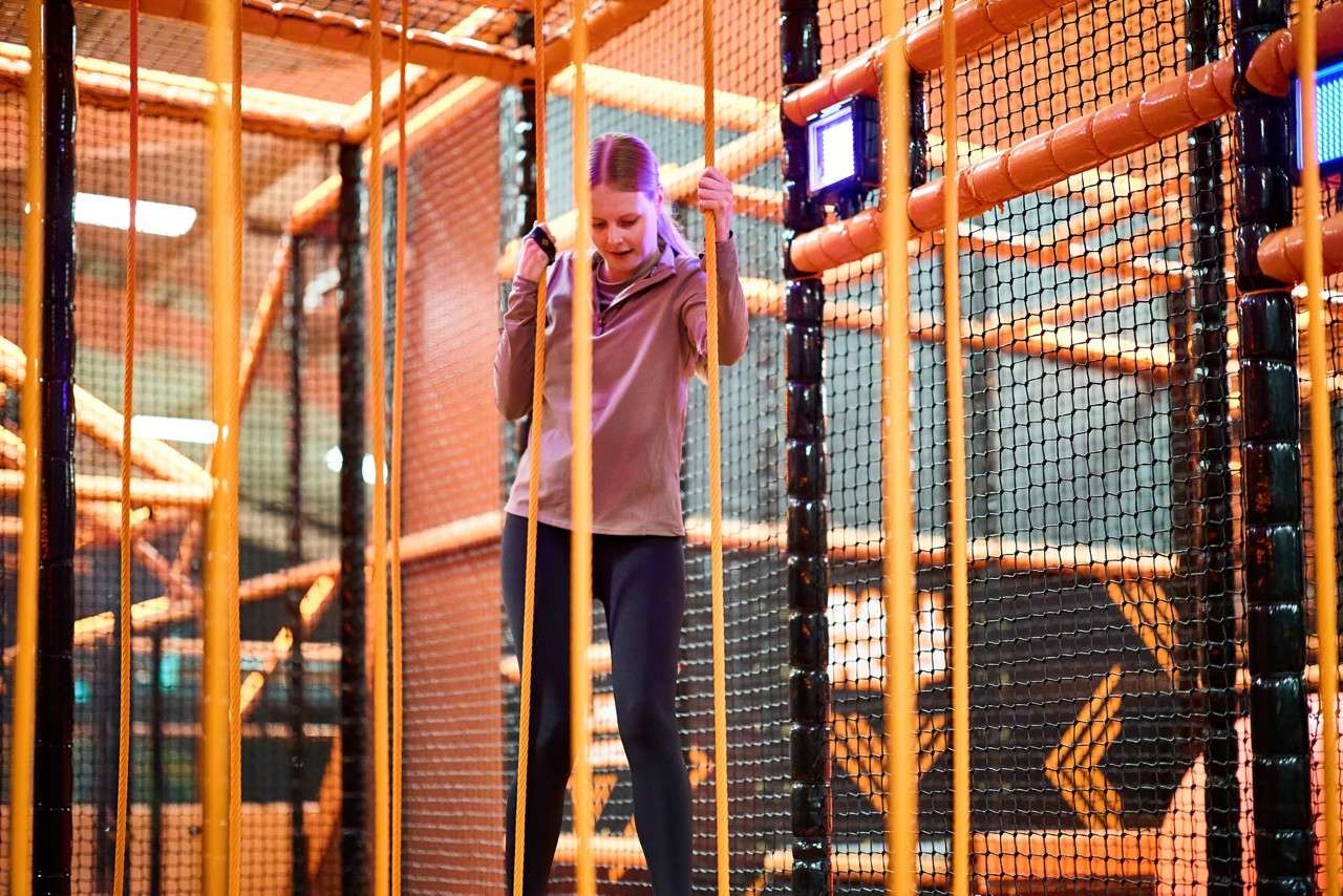 Person grips two vertical ropes, stepping across a narrow suspended bridge within an indoor obstacle course, surrounded by orange tubular frames, black safety netting, and small glowing lights.