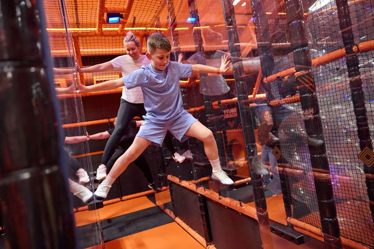 Boy spreads arms and legs to climb between two walls, pressing feet on panels. Behind him, children follow inside an orange, netted indoor obstacle course with padded platforms.