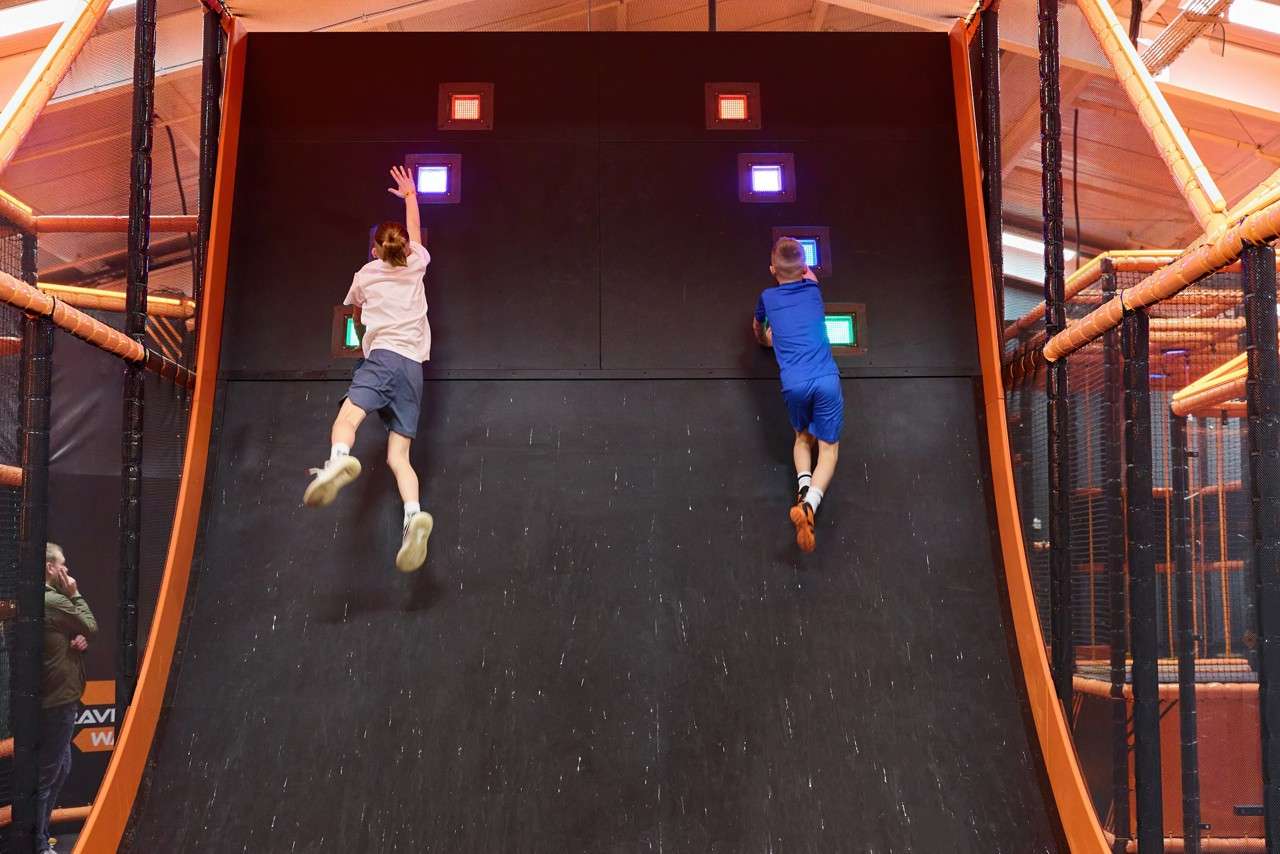 Two children sprint up a steep curved wall, reaching for glowing colored squares. They compete on an indoor obstacle course surrounded by orange-framed nets and beams under warehouse lighting.