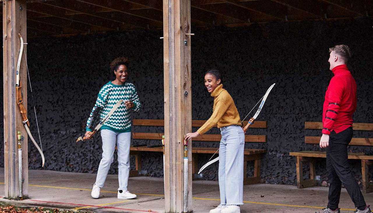 Three people with recurve bows chat and laugh between wooden posts, standing in a covered archery range with benches behind them; arrows and hanging bows rest nearby on the structure.