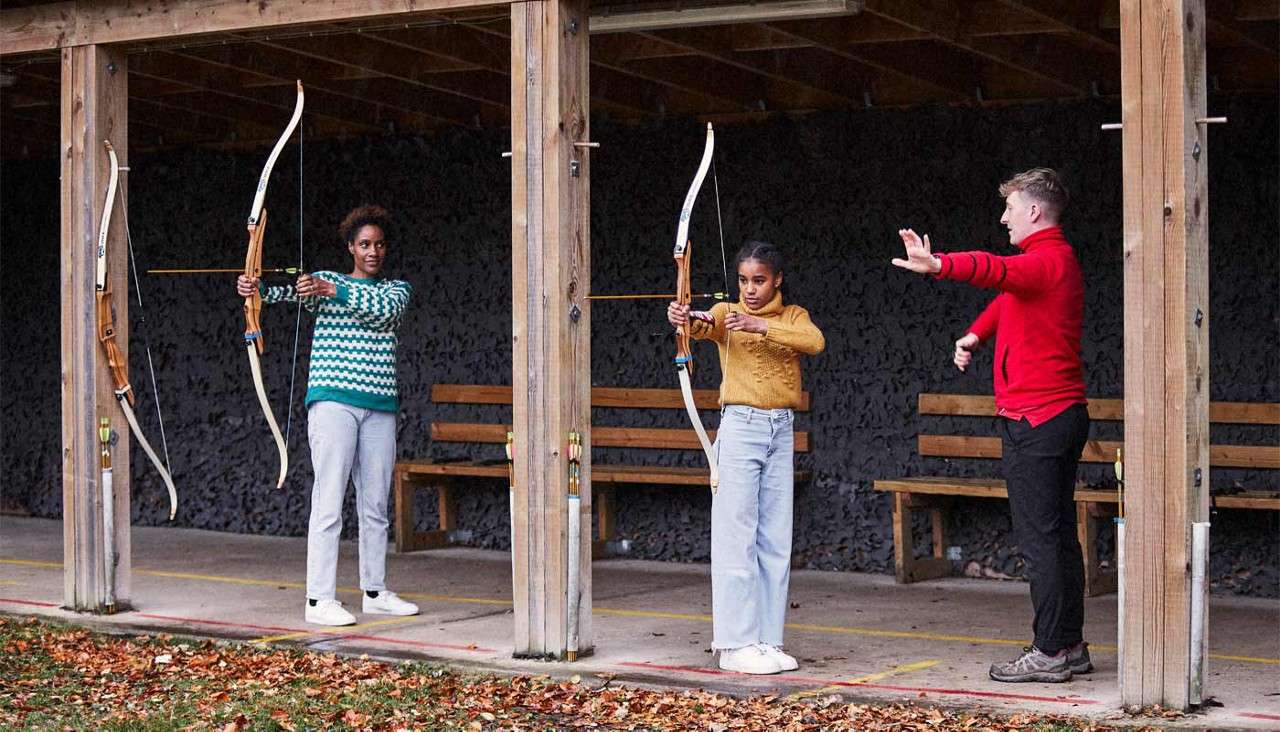 Two archers draw recurve bows while an instructor gestures guidance, standing under a wooden, covered archery lane with hanging spare bows, benches behind them, and fallen autumn leaves along the foreground.