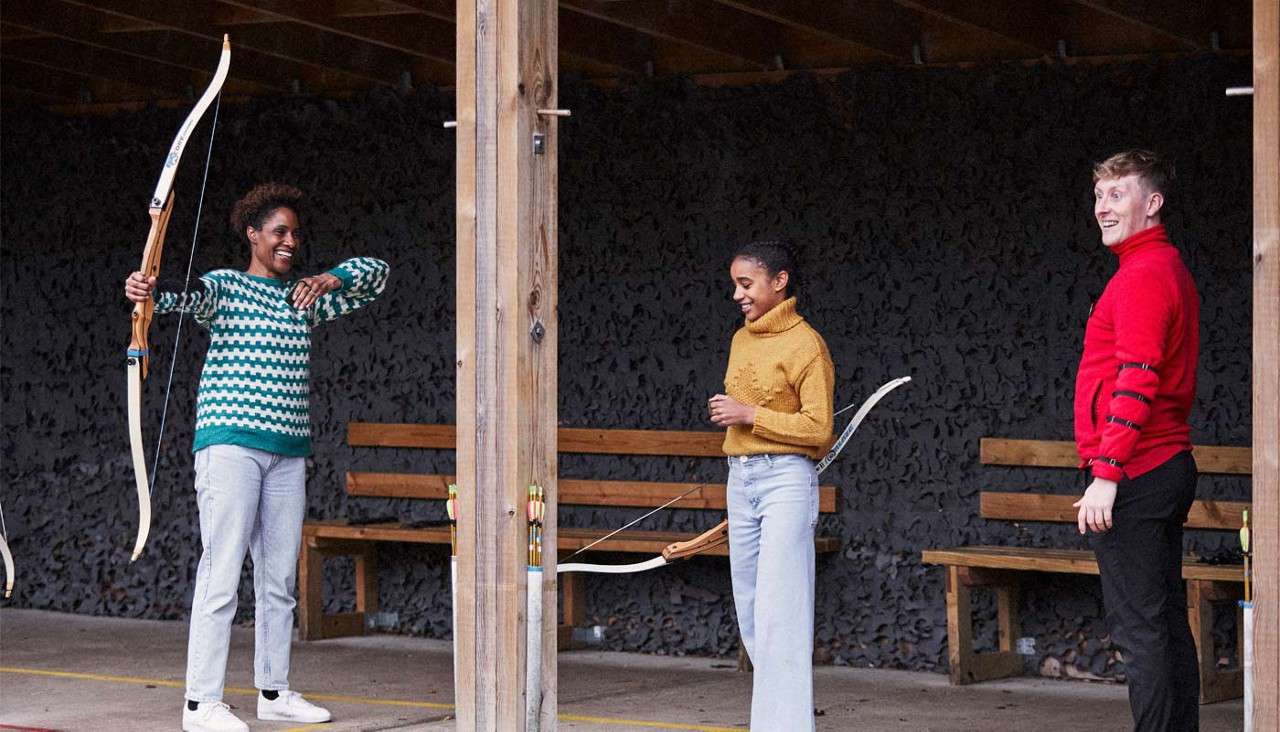 Archers practice at a covered range; three adults hold recurve bows, one drawing, others waiting and smiling; wooden posts, benches, arrow holders, and a dark backstop line the sheltered lane.
