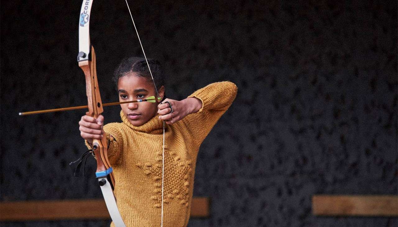 Young archer draws a recurve bow, aiming an arrow with focus; indoor range with dark wall background. Text: CORE on the bow.