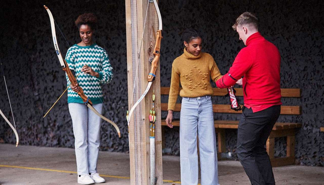 Archer receives guidance; instructor fastens an arm guard while another participant holds a recurve bow. Archery range with wooden posts, hanging bows, arrow holder, bench. Text: “CORE” on a bow limb.