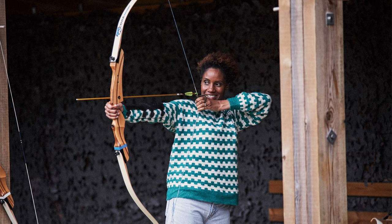 Archer draws a recurve bow and aims an arrow while smiling, inside a sheltered archery range with wooden posts and a dark backstop. Visible text: CORE.