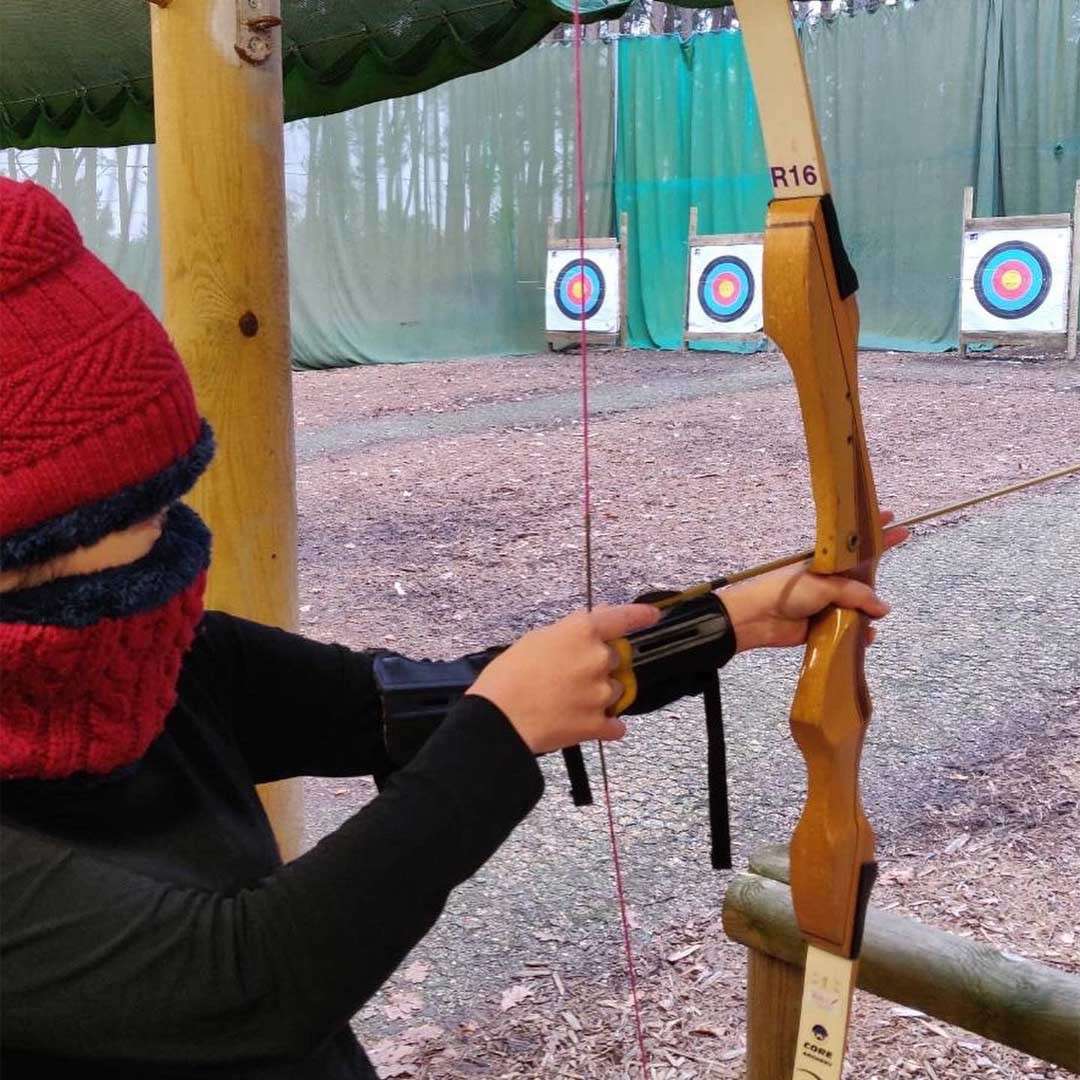Archer draws a recurve bow while aiming at distant circular targets; outdoor range with green safety nets and wooden posts. Visible text: R16, CORE.