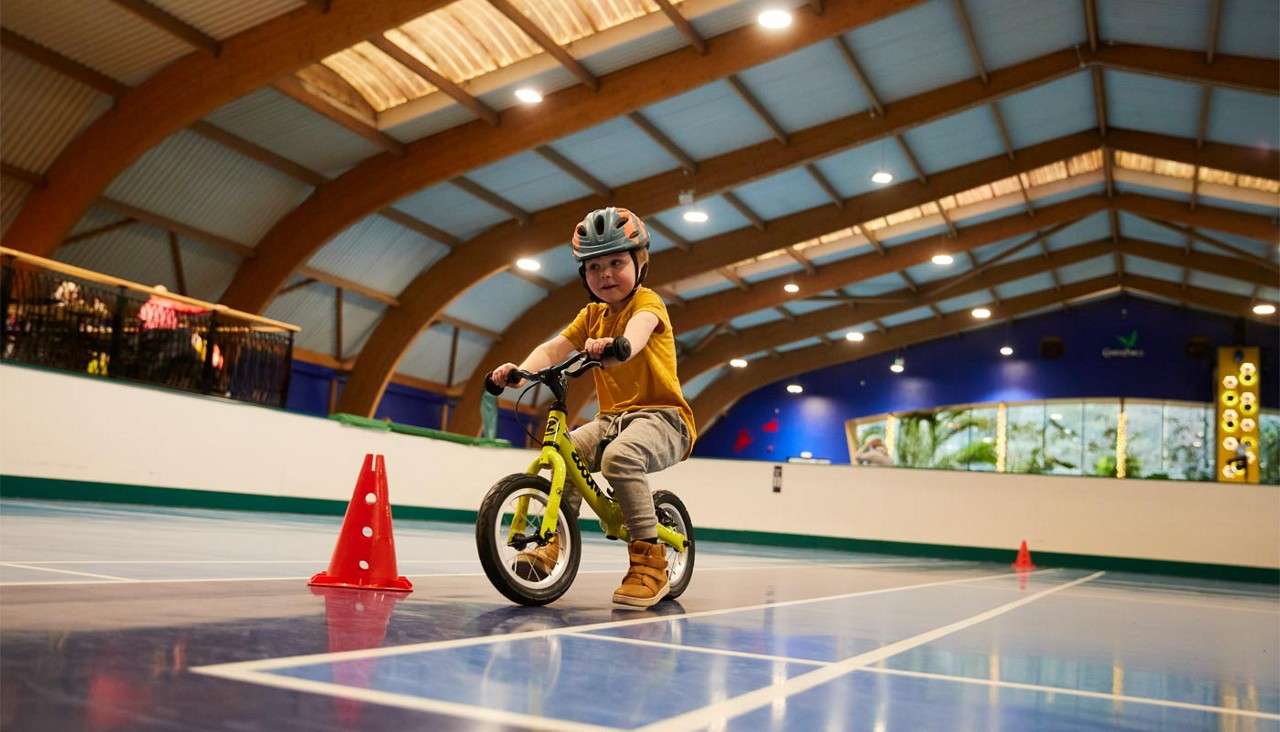 Child on a small balance bike navigates around an orange cone, wearing helmet and pads, inside a lit indoor sports hall with arched wooden beams and blue court lines.