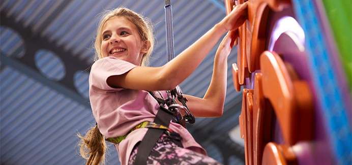 Child climber ascends a colorful indoor wall, smiling while clipped into a safety harness and auto-belay rope; metal ceiling and bright play-gym environment surround the activity.