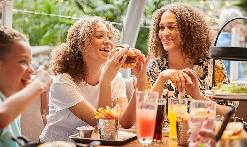 Young woman holds a burger and smiles before biting, seated with friends at a casual restaurant table filled with fries, drinks, and condiments, with greenery and string lights in the background.