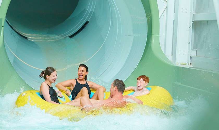 Four people on a yellow inflatable raft splash out of a enclosed waterslide, laughing and holding handles, surrounded by churning water inside an indoor water park with green tunnel walls.