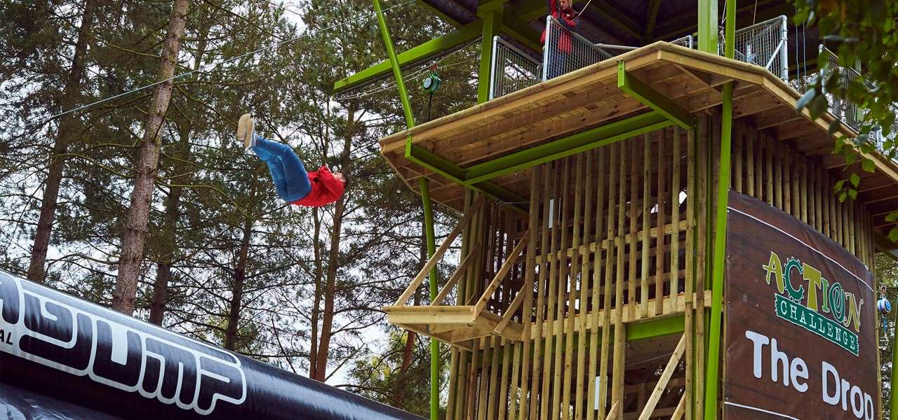 Person flips backward off a high wooden platform toward a large inflatable landing bag in a forest adventure park. Text visible: ACTION CHALLENGE; The Dro; JUMP.