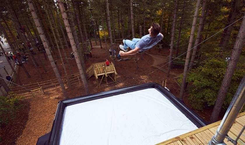 Person falls backward through the air, about to land on a large white airbag, within a treetop adventure park of wooden platforms and trees, with onlookers below.