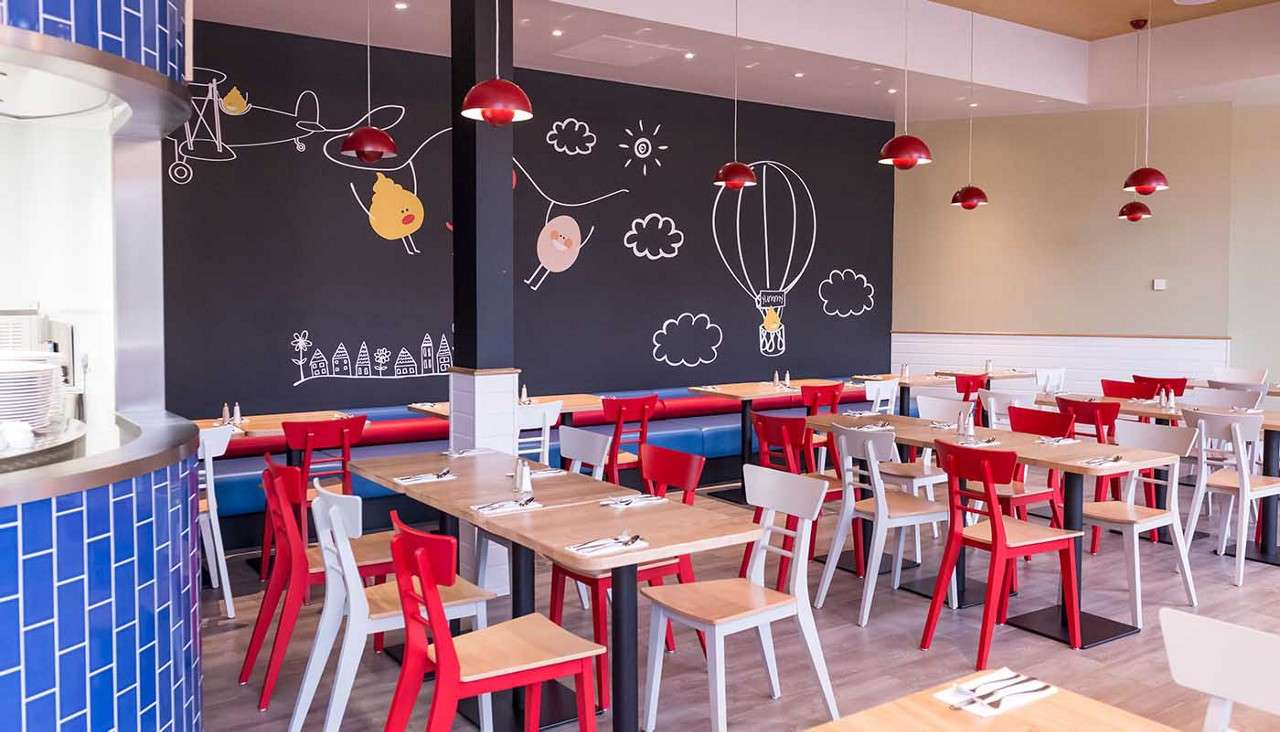 Colorful dining chairs rest around wooden tables, awaiting guests, in a family-friendly restaurant decorated with red pendant lights and a chalkboard-style mural of planes, clouds, and a hot-air balloon.