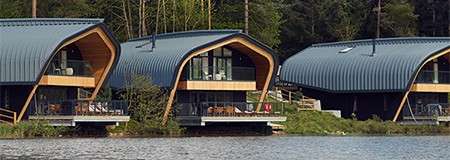 Curved-roof wooden cabins sit on stilts, overlooking a calm lake; balconies hold outdoor furniture, while trees and a forested shoreline surround the structures in a tranquil, natural setting.