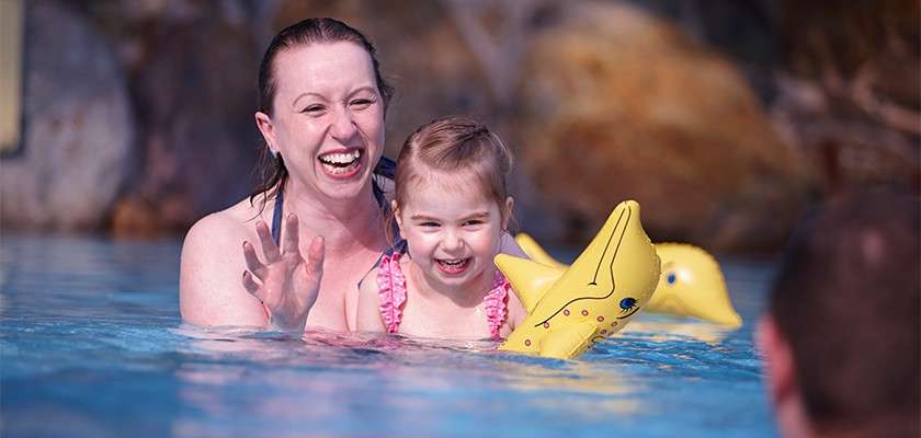Woman and young girl laugh while wading chest-deep, the child wearing yellow inflatable arm bands; they face a person in the foreground within a blue swimming pool with rocky backdrop.