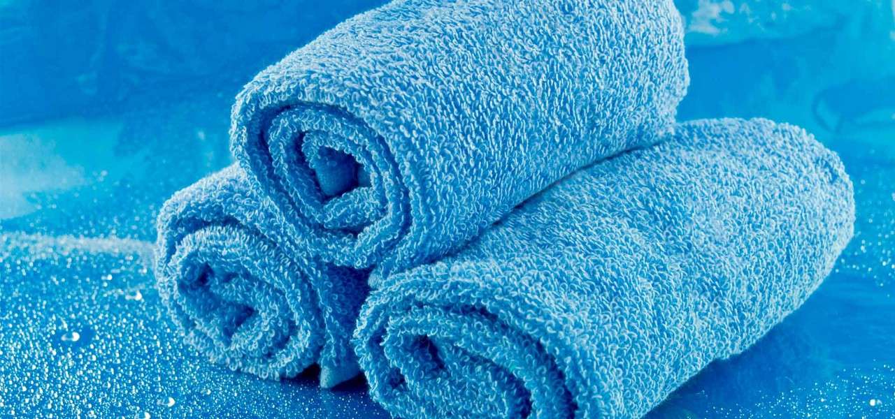 Three rolled blue towels rest stacked on a damp surface, beads of water glistening around them, in a cool-toned, spa-like setting.