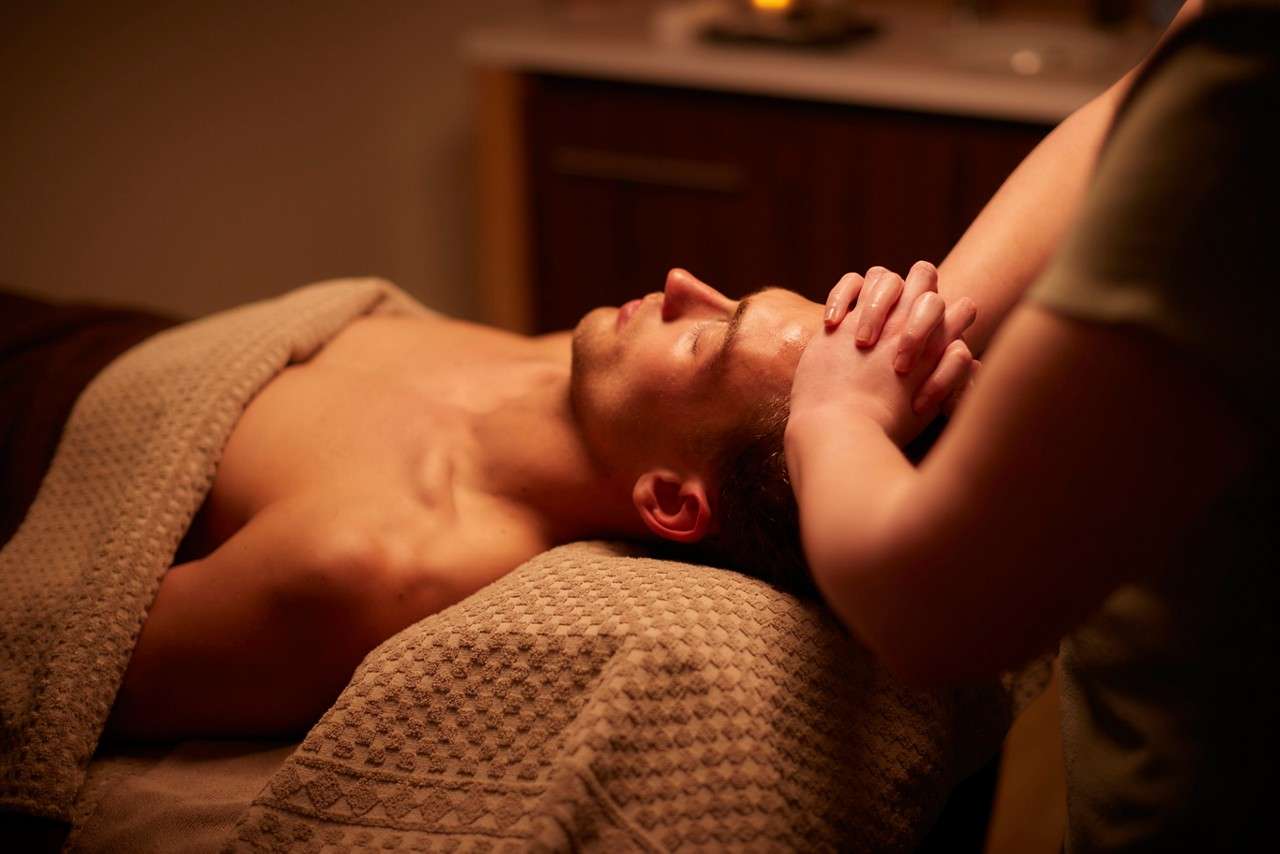 Person lies on a massage table, eyes closed, receiving a head massage from a therapist, in a dimly lit spa room with warm tones and a textured towel and blanket.