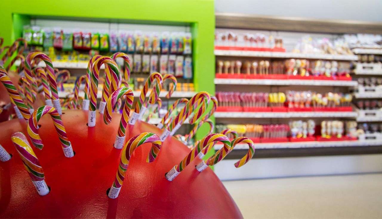 Striped candy canes stand upright, inserted into a red domed display. Behind them, a bright candy shop with a green wall shows shelves of assorted lollipops and sweets, blurred.