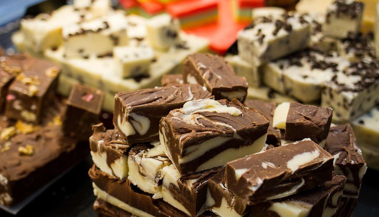 Swirled chocolate-and-vanilla fudge pieces stack in a heap, foreground, while other assorted fudge blocks rest on trays in the background at a confectionery display.