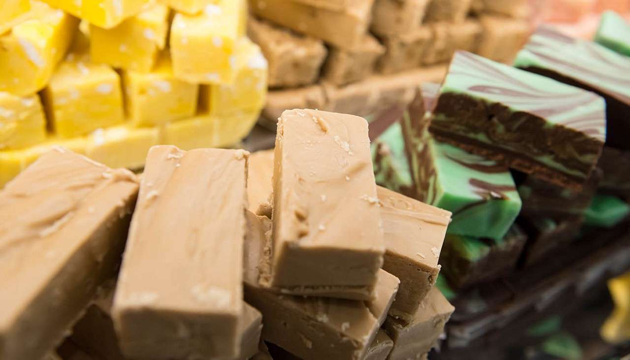 Fudge pieces stack into tan bars, resting in a neat pile; surrounding context shows additional varieties—yellow cubes and green-brown marbled slabs—on a confectionery display, likely a sweet shop counter.