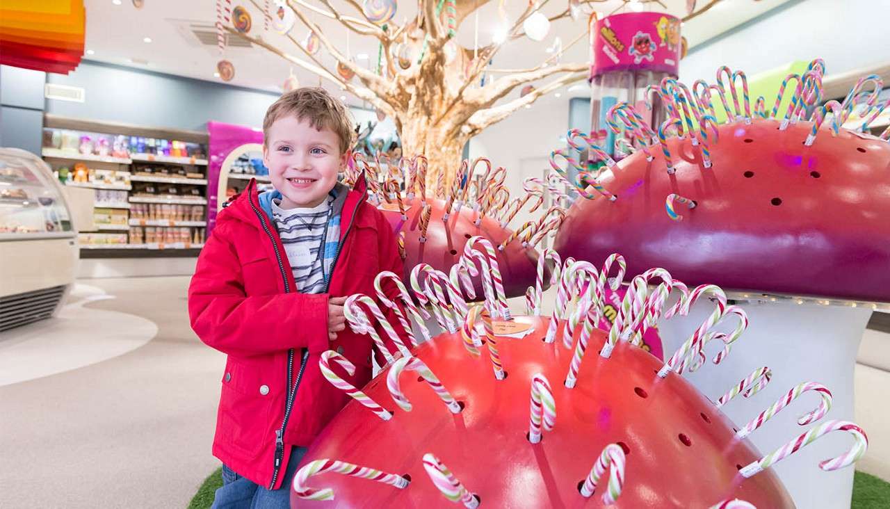 Child smiles while standing beside large red mushroom-shaped displays holding striped candy canes, in a bright, whimsical candy store with a golden candy tree centerpiece and shelves of sweets behind.