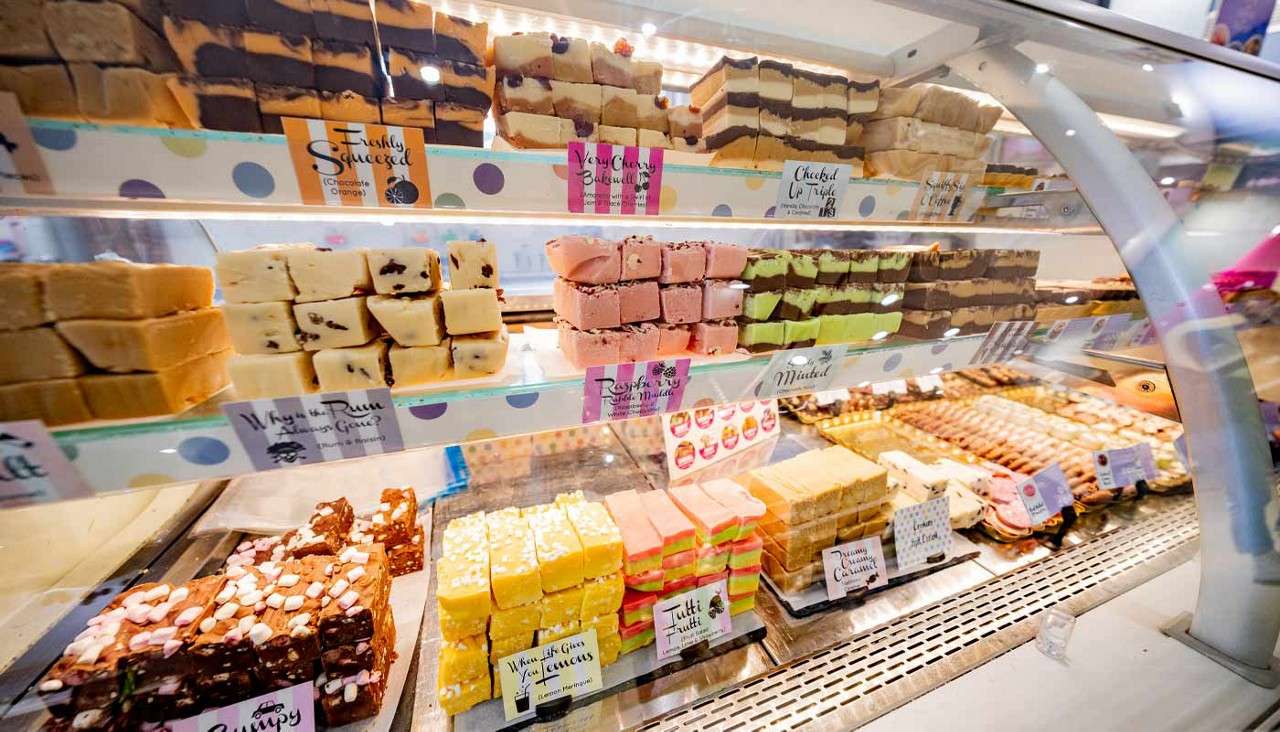 Stacks of colorful fudge cubes sit neatly arranged on shelves inside a glass display case at a sweets shop.

Text: Freshly Squeezed; Raspberry; Minted; Tutti Frutti; When Life Gives You Lemons; Dreamy Creamy Coconut.