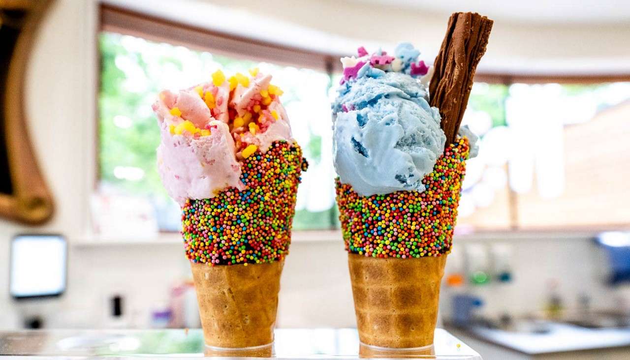 Two sprinkle-coated waffle cones hold pink and blue ice cream, topped with candies; the right includes a chocolate flake. They stand on a counter inside a bright ice-cream shop.