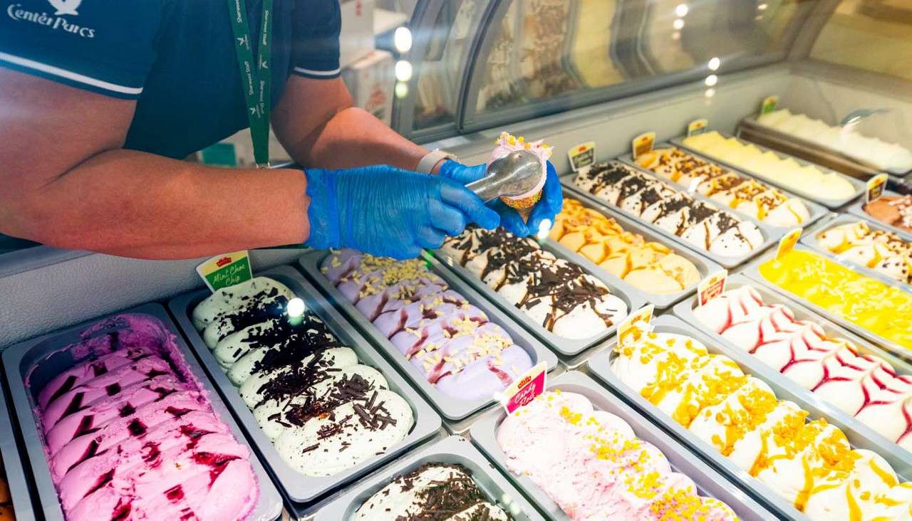 Server scoops ice cream into a cone, over a display case of colorful gelato trays with toppings inside a shop.
Text: Center Parcs; Mint Choc Chip.