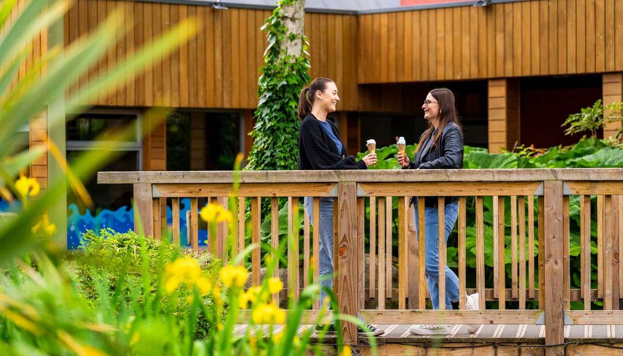 Two people hold ice cream cones and chat on a wooden footbridge. Green plants and yellow flowers surround them, with a timber building and ivy-wrapped tree in the background.