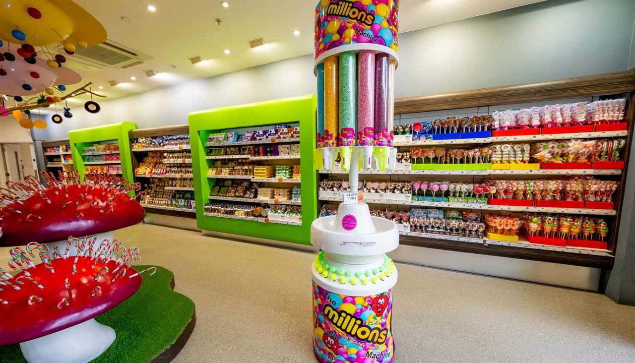 Candy dispenser displays multi-colored pellets, standing in a bright candy store with lollipop-lined shelves, green-framed displays, and whimsical red mushroom stands. Text: millions; Machine.