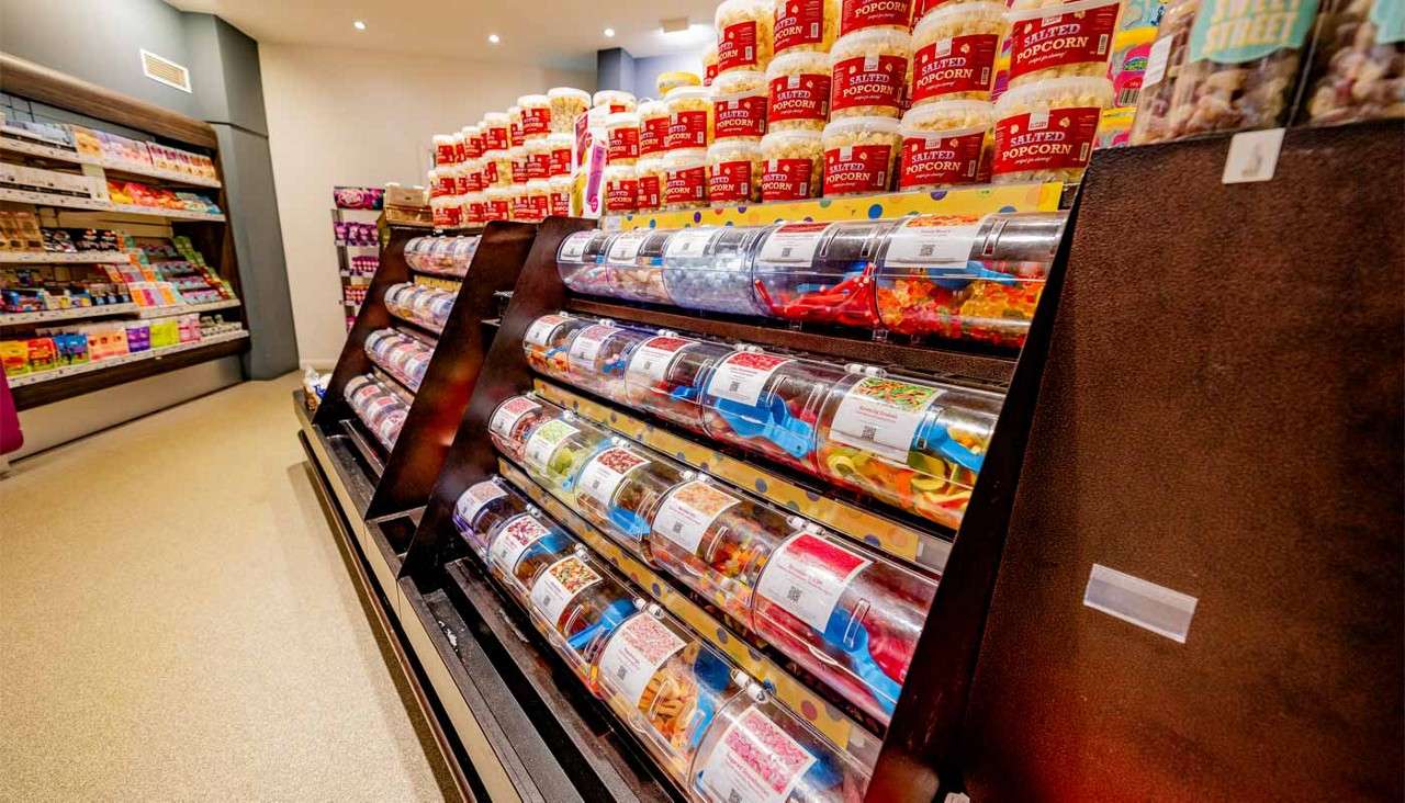 Clear cylindrical candy bins display assorted gummies with scoops on angled racks; behind them, stacked tubs read “SALTED POPCORN”; left shelves hold packaged sweets in a brightly lit shop.