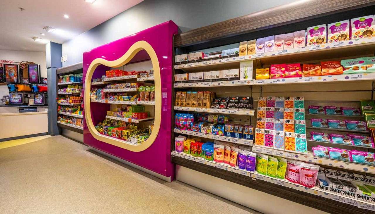 Snack shelves display assorted candies and snacks, lining a bright store aisle with a magenta rounded frame and a drinks counter on the left. Text: HIP; SMAX; VEGAN FUDGE. Additional small labels unreadable.