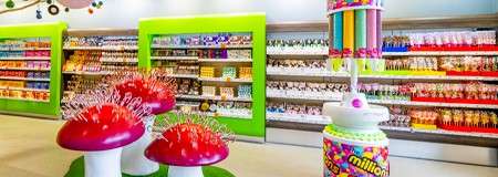 Mushroom-shaped lollipop displays stand prominently; rows of colorful packaged sweets line shelves in a bright candy store. Text: “millions” printed on a tall cylindrical dispenser.