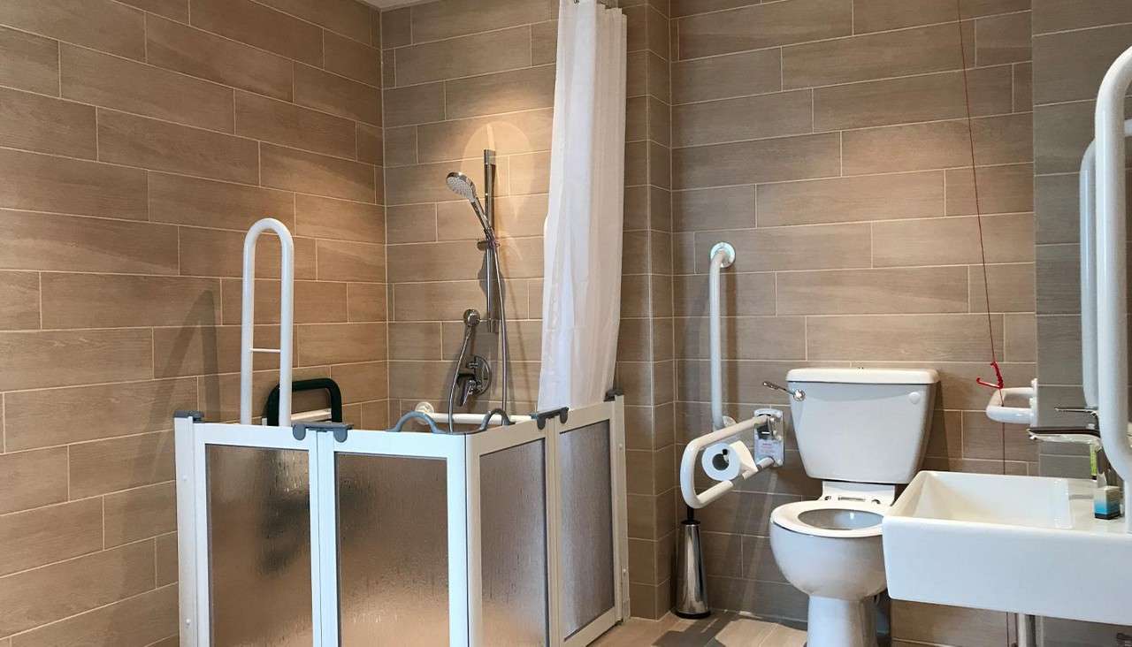 Accessible bathroom features—shower area with handheld head and half-height folding screens; toilet with grab bars and paper holder; wall-mounted sink—set within beige tiled room, curtain and emergency pull cord.