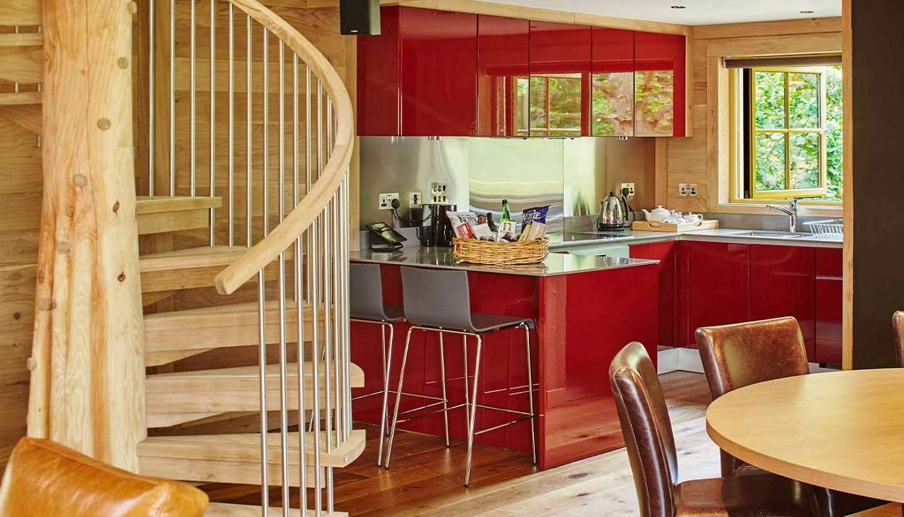 Spiral wooden staircase curves upward beside a kitchen; glossy red cabinets and steel counters frame bar stools and a basket, with window, sink, round dining table, and leather chairs.