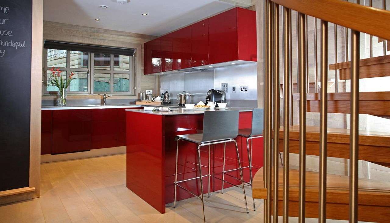 Red kitchen island hosts two stools and breakfast setup; glossy red cabinets and stainless backsplash surround it, with a window, vase of flowers, and staircase railing framing the modern kitchen.