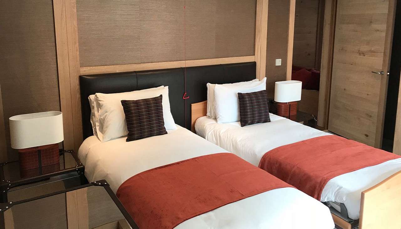 Twin beds sit neatly made with white linens, orange throws, and striped pillows, flanked by lamps on glass nightstands, within a wood-paneled room with doors and a ceiling pull cord.