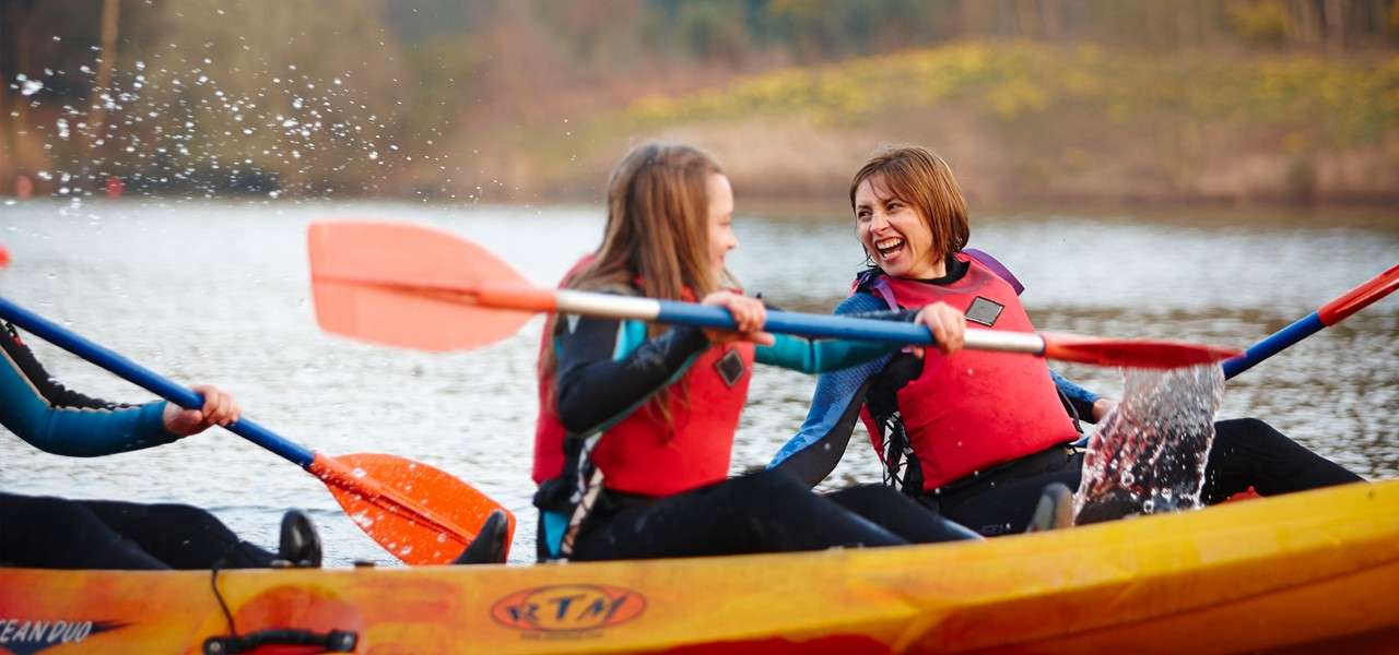 Two kayakers splash each other with orange paddles, laughing. They sit in a yellow tandem kayak on a calm lake with forested shore. Text: “RTM”, “OCEAN DUO”.