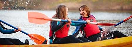Two kayakers paddle and splash water, smiling and facing each other, wearing red life jackets in a yellow kayak on a calm lake with a blurred wooded shoreline.