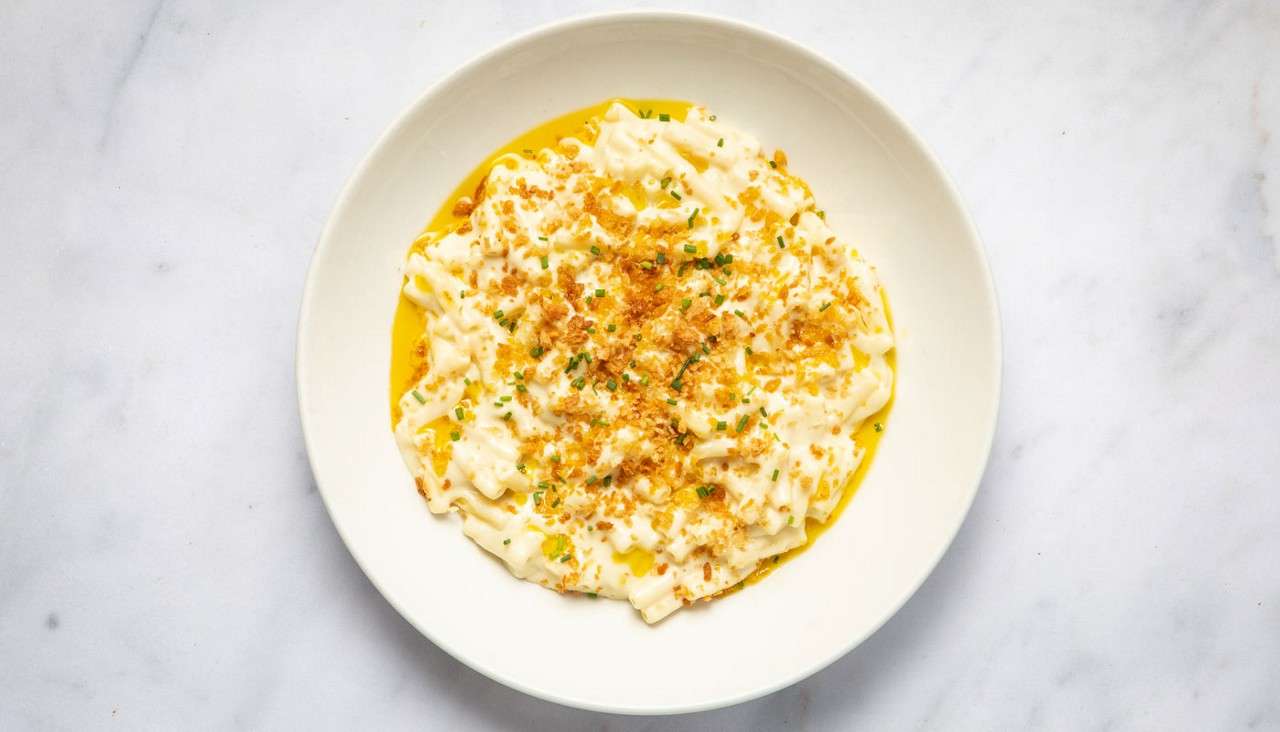 Creamy pasta rests topped with golden breadcrumbs and chopped chives, pooling with butter sauce, on a white plate centered on a light marble tabletop.
