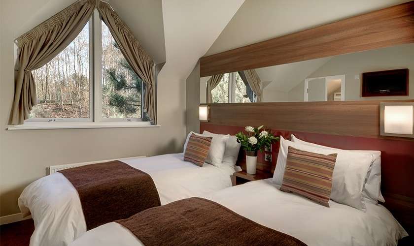 Twin beds sit neatly with brown throws and striped pillows in a sloped-ceiling room, with a large curtained window, long wall mirror, sconces, bedside flowers, and trees outside.