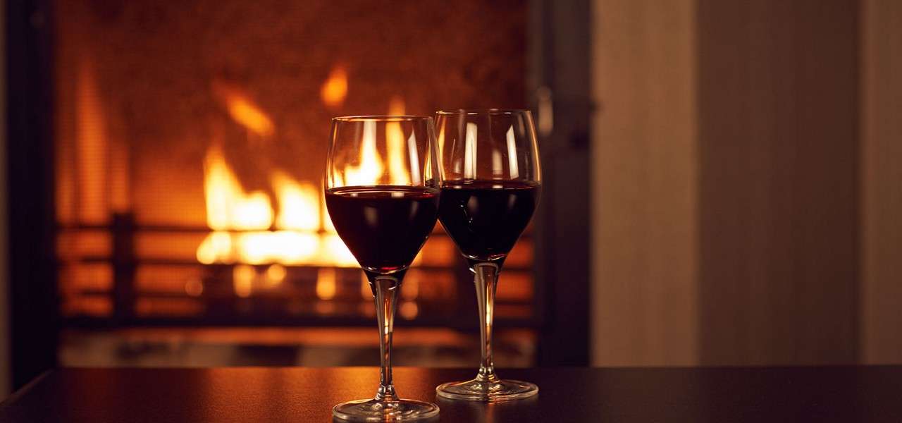 Two red-wine glasses stand side by side, reflecting firelight, on a dark tabletop; behind them, a lit fireplace glows warmly in a cozy indoor room.