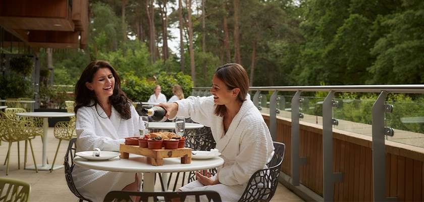 Two people in white bathrobes share lunch; one pours a drink into a glass while they sit at a round table on an outdoor terrace overlooking trees and other diners.