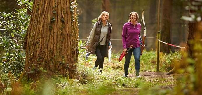 Two adults walk along a wooded trail, carrying archery gear—bows and arrows—amid tall trees, green undergrowth, and a red-and-white ribbon marking the course.