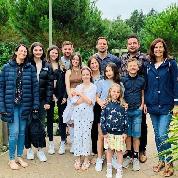 Group of thirteen people pose and smile, arranged in two rows. Adults and children wear casual clothes and jackets. Paved path bordered by greenery and pine trees, overcast day.