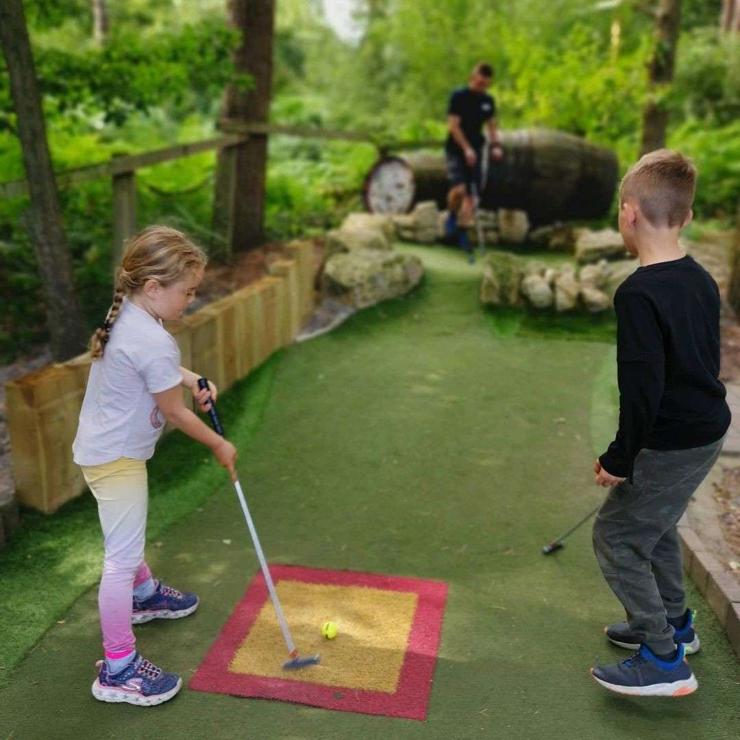 Girl grips a putter, ready to hit a yellow ball on a tee square; a boy watches as others play near rocks and a barrel on a forested mini-golf course.